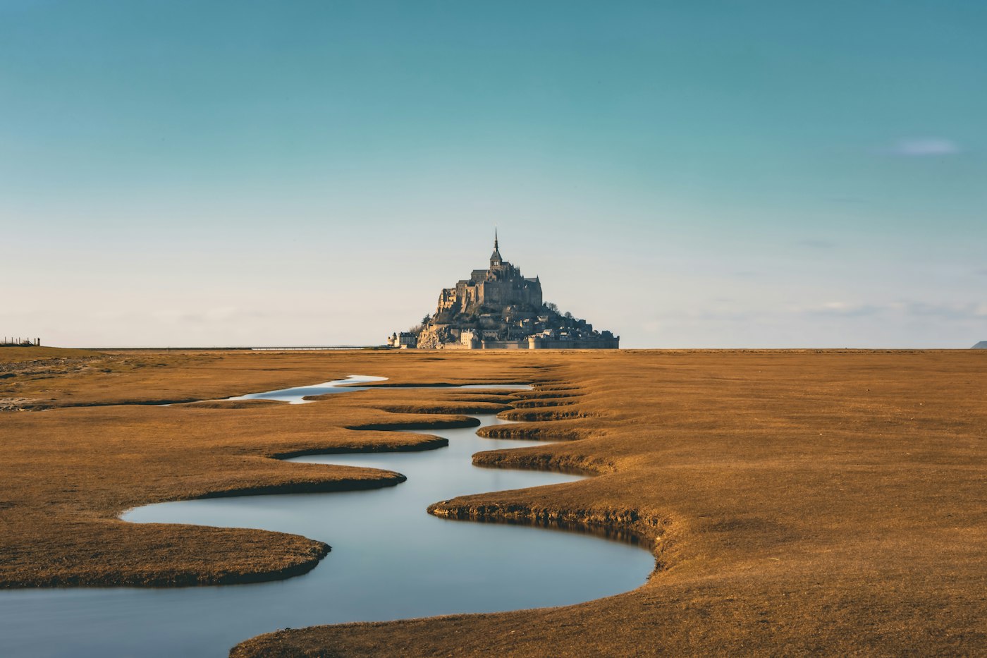 A distant view of Mont-Saint-Michel across golden tidal marshes and a winding water channel